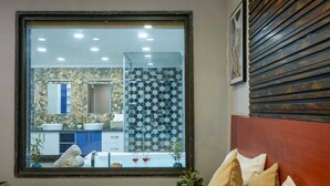 Une chambre avec une grande fenêtre, un lit avec des oreillers, une table de chevet et une vue sur une salle de bain dont le mur derrière l’évier est recouvert de carreaux.