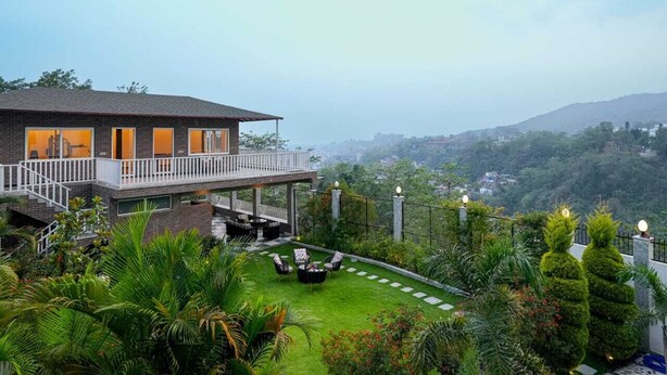Un bâtiment à deux étages doté d’un balcon, d’un jardin avec une pelouse et des plantes en pot, et offrant une vue sur un paysage vallonné.