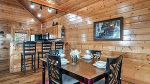 Interior de cabana de madeira com mesa de jantar posta para quatro, paredes de madeira, área de cozinha e quadro com fotografia de animais selvagens.