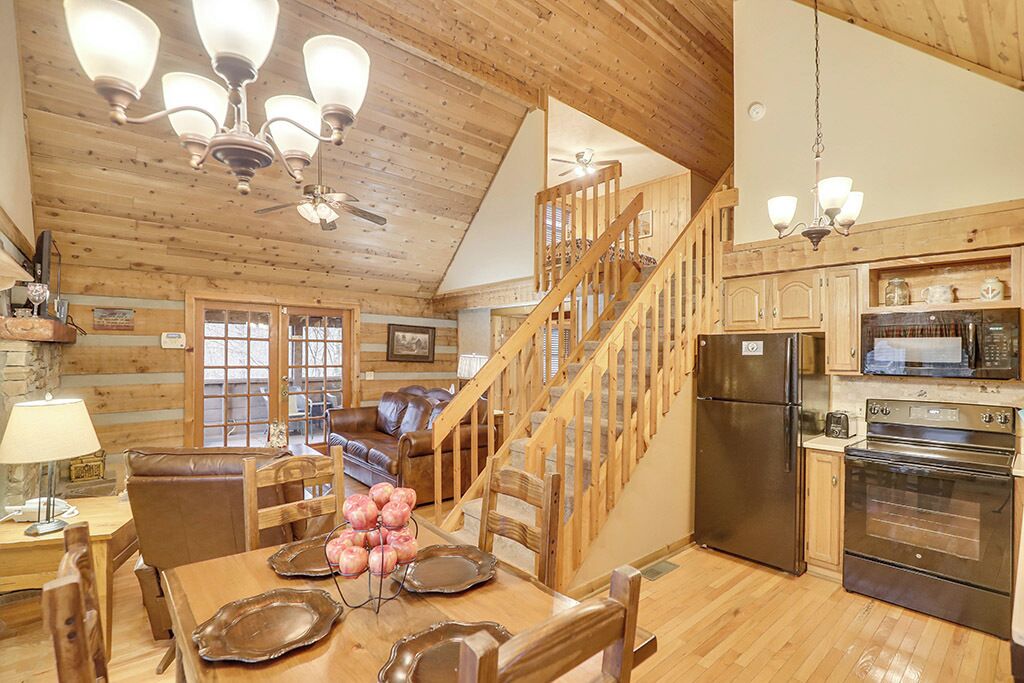 A cozy cabin interior with a dining area, wooden staircase, kitchen, and a fireplace.