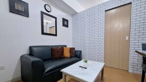 A room with a black leather sofa, a white coffee table, a clock, a mirror, and a door with a brick wall. - Homey 1BR with Balcony City View in Grace Res (Taguig City)