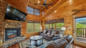 A cozy cabin interior with a stone fireplace, leather seating, a flat-screen TV, and large windows offering a scenic view.