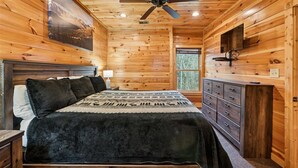 A wooden cabin bedroom with a bed, dresser, ceiling fan, and a large window.