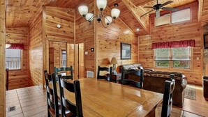 Ein Holzblockhaus-Interieur mit Esstisch, Stühlen, einer Couch, einem Deckenventilator und einer Chandelier.