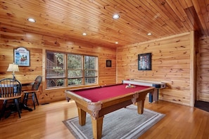 L’intérieur d’un chalet en bois comprend une table de billard, des chaises et une fenêtre donnant sur la forêt.