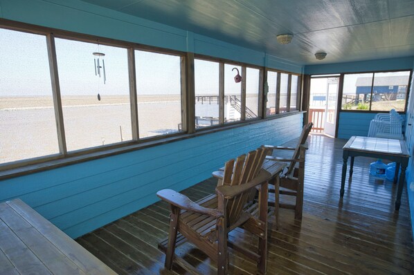 A covered deck with wooden furniture, large windows overlooking the beach, and a view of the ocean.