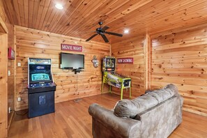 Una habitación acogedora con paredes de madera, ventilador de techo, sofá y juegos de arcade, incluyendo una máquina temática de Shrek.