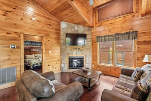 A log cabin interior with a stone fireplace, wooden walls, a television mounted above it, and a cozy seating area with a sofa and armchairs. - Romantic Cabin w/ Hot Tub, Games & Fireplace (Gatlinburg)