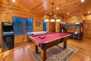 Une table de billard recouverte d’un feutre rouge, une table de baby-foot et une table de ping-pong se trouvent dans une pièce aux murs et au plafond en bois.
