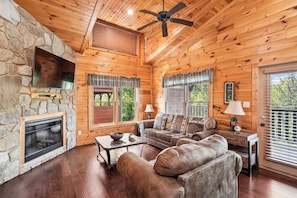 A log cabin interior with a stone fireplace, wooden walls, a ceiling fan, and a leather sofa. - Romantic Cabin w/ Hot Tub, Games & Fireplace (Gatlinburg)