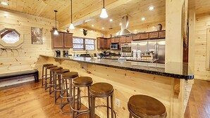 A wooden cabin interior with a kitchen, bar stools, and a large window. - Expansive Luxury Cabin w/ Games, Theater & Views (Gatlinburg)