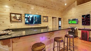 A wooden interior with a bar area featuring stools, a television, and a gaming console. - Expansive Luxury Cabin w/ Games, Theater & Views (Gatlinburg)