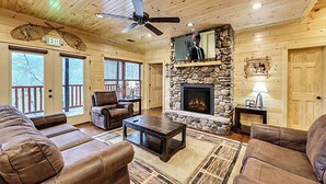 A cozy cabin interior with a stone fireplace, leather furniture, and a ceiling fan. - Expansive Luxury Cabin w/ Games, Theater & Views (Gatlinburg)