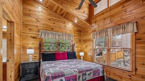 Ein Schlafzimmer in einer Blockhütte mit einem Bett, zwei Nachttischen, einem Fenster mit Vorhängen und Blick ins Freie.