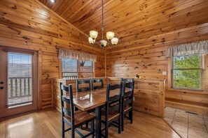 Interior de una cabaña de madera con una mesa de comedor, sillas, una araña y ventanas.