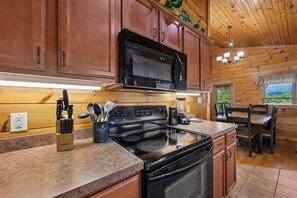 A kitchen with wooden cabinets, a black microwave, a stove, and a dining area with a table and chairs. - Modern Mountain Getaway w/ Hot Tub & Arcade (Gatlinburg)