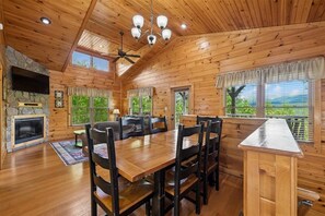 A wooden cabin interior with a dining area, fireplace, and large windows offering a view. - Modern Mountain Getaway w/ Hot Tub & Arcade (Gatlinburg)