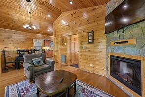 A cozy cabin interior with a fireplace, wooden walls, a dining area, and a living room with a sofa and coffee table. - Modern Mountain Getaway w/ Hot Tub & Arcade (Gatlinburg)