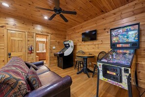A room with a wooden interior, a ceiling fan, a television mounted on the wall, a sofa with patterned cushions, and a gaming machine. - Modern Mountain Getaway w/ Hot Tub & Arcade (Gatlinburg)
