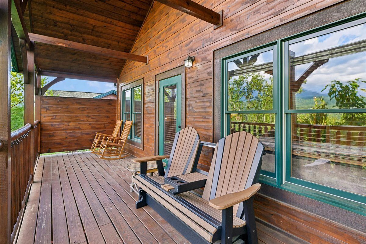 Une terrasse en bois avec des fauteuils Adirondack et un banc, de grandes fenêtres aux cadres verts et une vue sur la verdure.