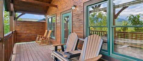 Una terraza de madera con sillas Adirondack y un banco, ventanas grandes con marcos verdes y vista a la vegetación.