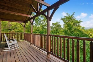 A wooden deck with a railing, a rocking chair, and a view of a forested area. - Modern Mountain Getaway w/ Hot Tub & Arcade (Gatlinburg)