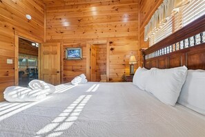 A wooden cabin bedroom with a large bed, a painting on the wall, and a window with curtains. - Modern Mountain Getaway w/ Hot Tub & Arcade (Gatlinburg)