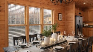 Une salle à manger aux murs lambrissés, avec une longue table dressée pour six personnes, un lustre et une vue dégagée sur l’extérieur grâce à de grandes fenêtres.