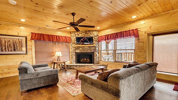 A cozy cabin interior with a stone fireplace, wooden walls, and a ceiling fan.