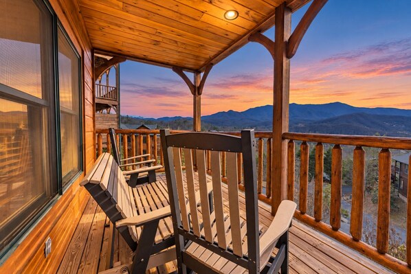 A wooden deck with two Adirondack chairs, a glass door, and a view of mountains.