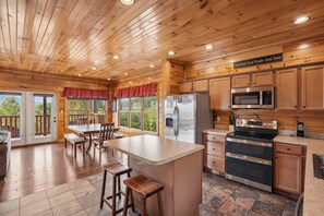 A wooden cabin interior with a kitchen, dining area, and a view through the windows.