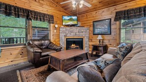 A cozy cabin interior with a stone fireplace, leather furniture, and a television mounted on the wall.
