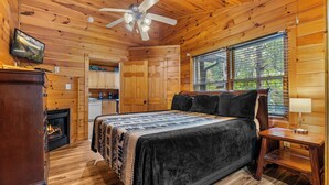 A cozy log cabin bedroom with a bed, a fireplace, a ceiling fan, and a view of the outdoors through the window. - Gatlinburg - A Creekside Cottage - 1 Bdrms. (Gatlinburg)