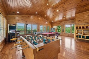 A wooden-floored room with a foosball table, pool table, and a bar area.