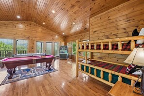 A wooden interior with a pool table, bunk beds, and a pool table with a blue and white patterned rug.