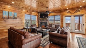 A log cabin interior with a stone fireplace, leather sofas, a flat-screen TV, and large windows.