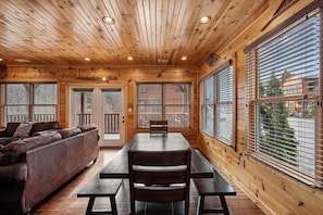 A wooden cabin interior with a sofa, dining table, and benches, large windows with blinds, and a view of the outside.