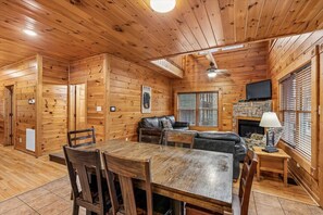 Interior de uma cabana de madeira com mesa de jantar, cadeiras, sofá, lareira e televisão.