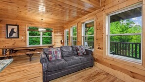 Interior de uma cabana de madeira com sofá de couro, janelas amplas e uma fotografia de urso na parede.