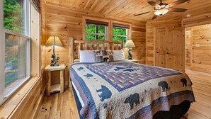 Quarto de cabana de madeira com uma cama, duas mesas de cabeceira, um ventilador de teto e janelas.