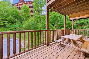 Une terrasse en bois, dotée d’une table de pique-nique et d’une rambarde, qui offre une vue sur une forêt verdoyante et un bâtiment avec des balcons.