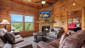 L’intérieur chaleureux d’un chalet avec une cheminée, des canapés en cuir, un téléviseur et de grandes fenêtres offrant une vue panoramique.