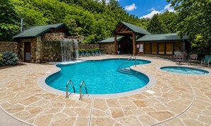 A pool area with two hot tubs, a waterfall feature, and stone-tiled flooring.