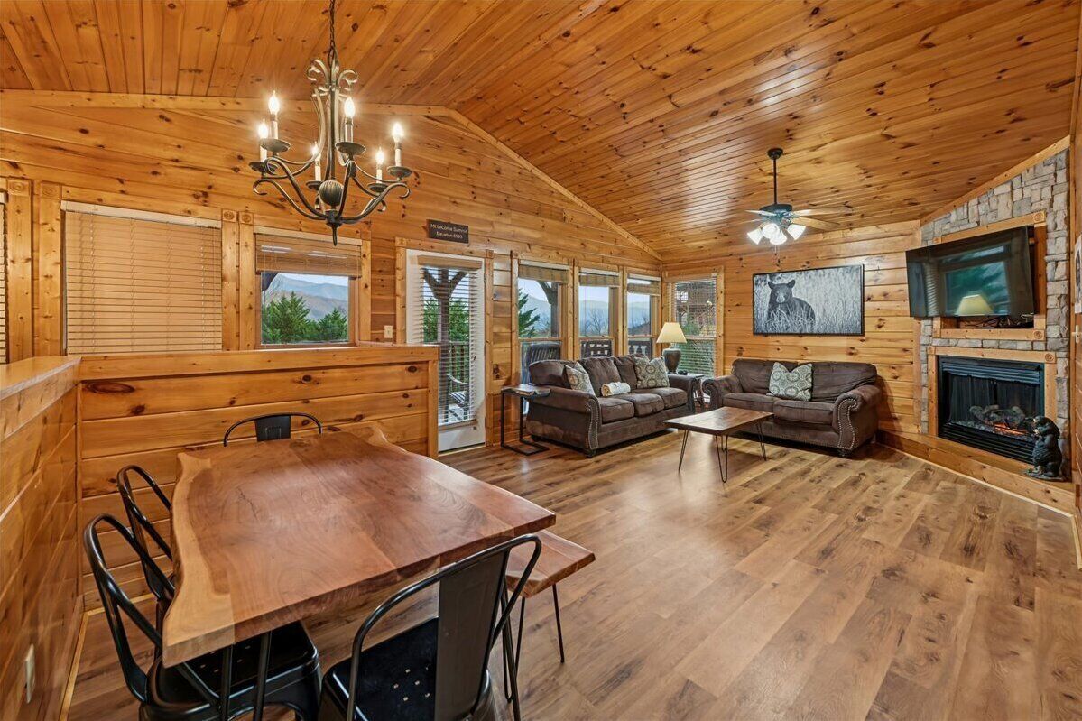 A log cabin interior with a dining area, sofas, a fireplace, and a large window overlooking greenery.