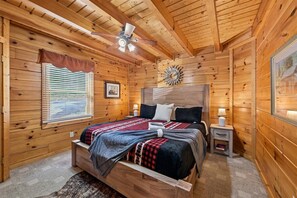 Een houten slaapkamer met een bed, nachtkastjes, een raam met zonwering, een plafondventilator en een wandklok.