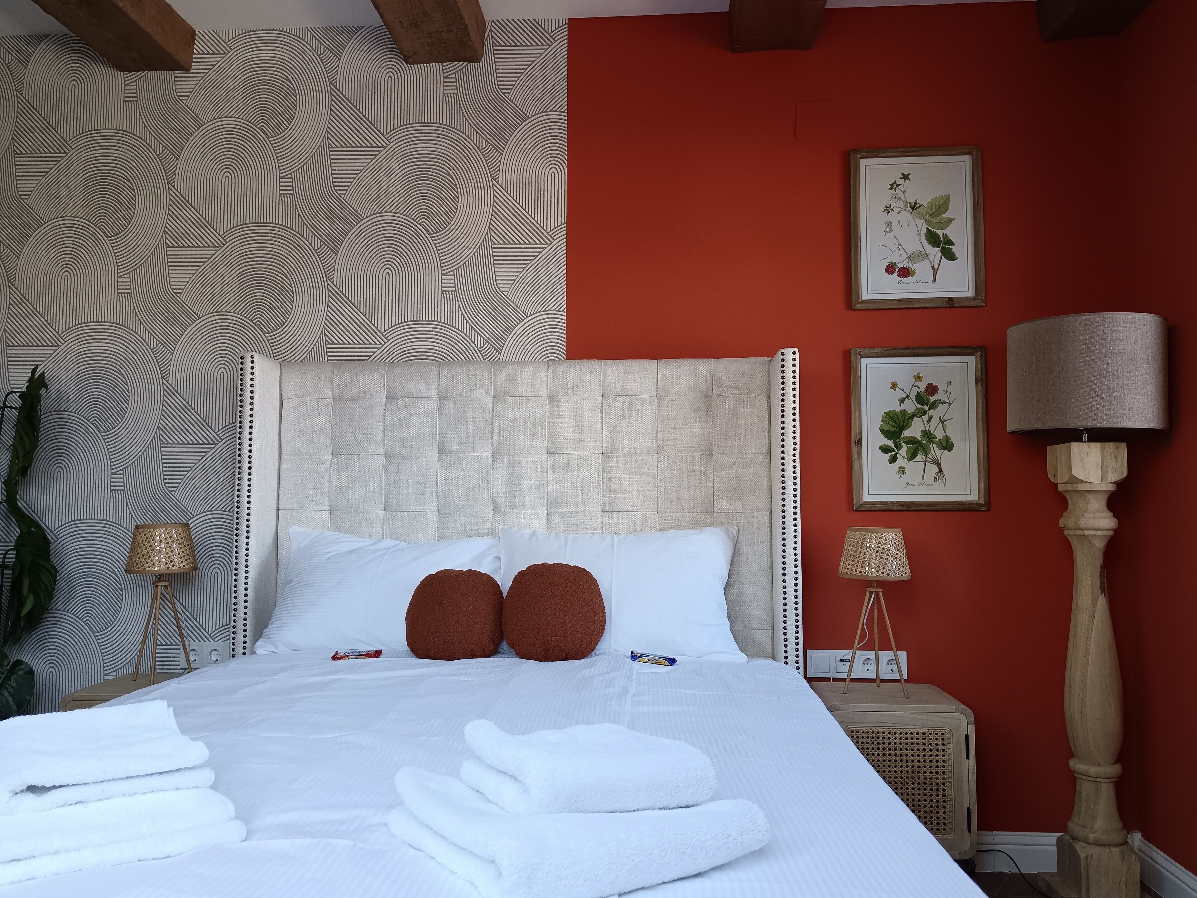A neatly made bed with white linens and two brown pillows, a red wall, and two framed botanical illustrations.