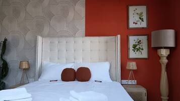 A neatly made bed with white linens and two brown pillows, a red wall, and two framed botanical illustrations.