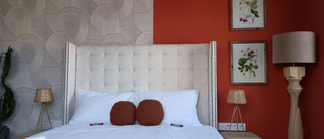 Una cama bien hecha con sábanas blancas y dos almohadas marrones, una pared roja y dos ilustraciones botánicas enmarcadas.