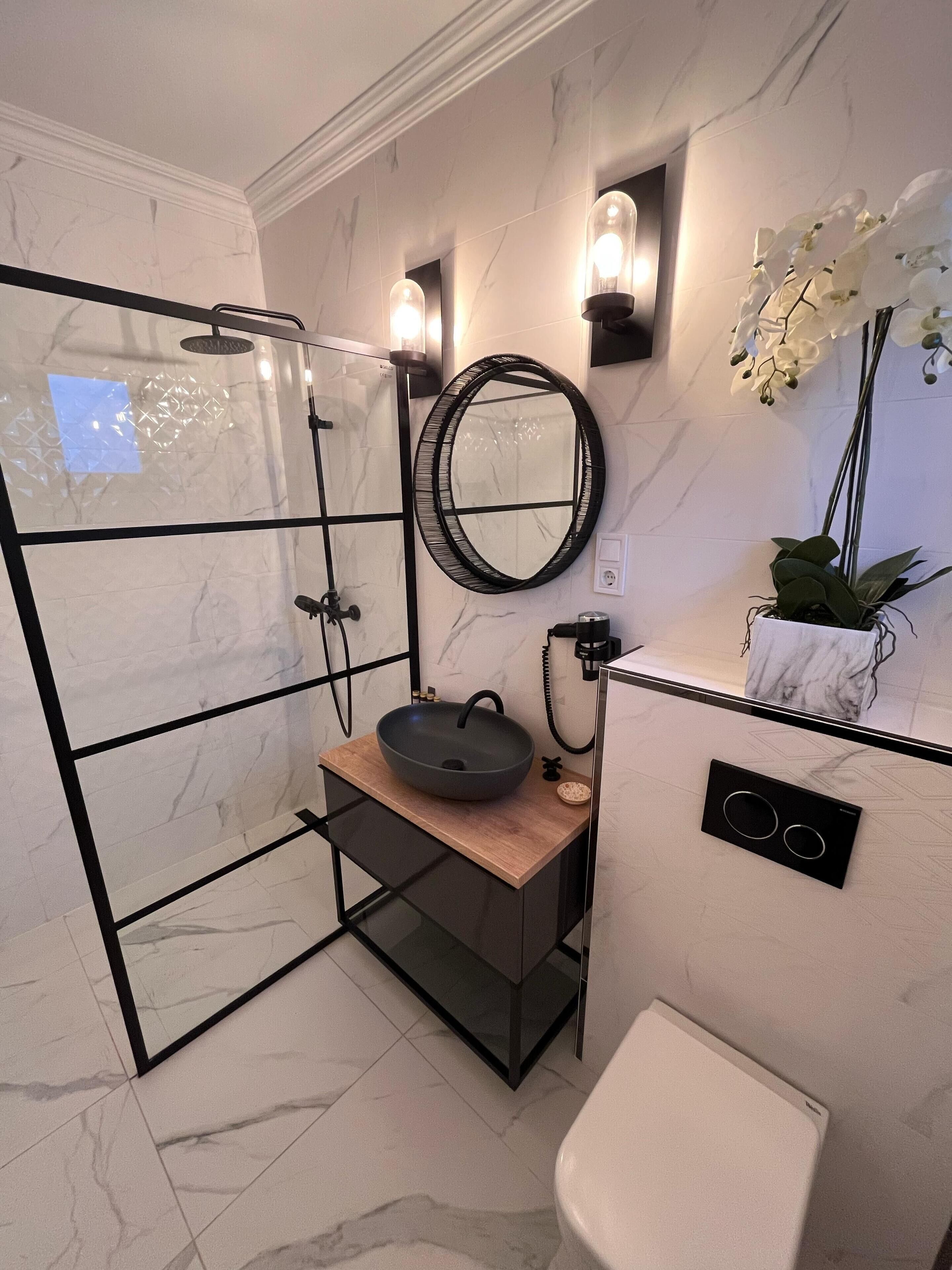 A modern bathroom with a glass shower enclosure, a round mirror, a vessel sink, and a potted orchid.