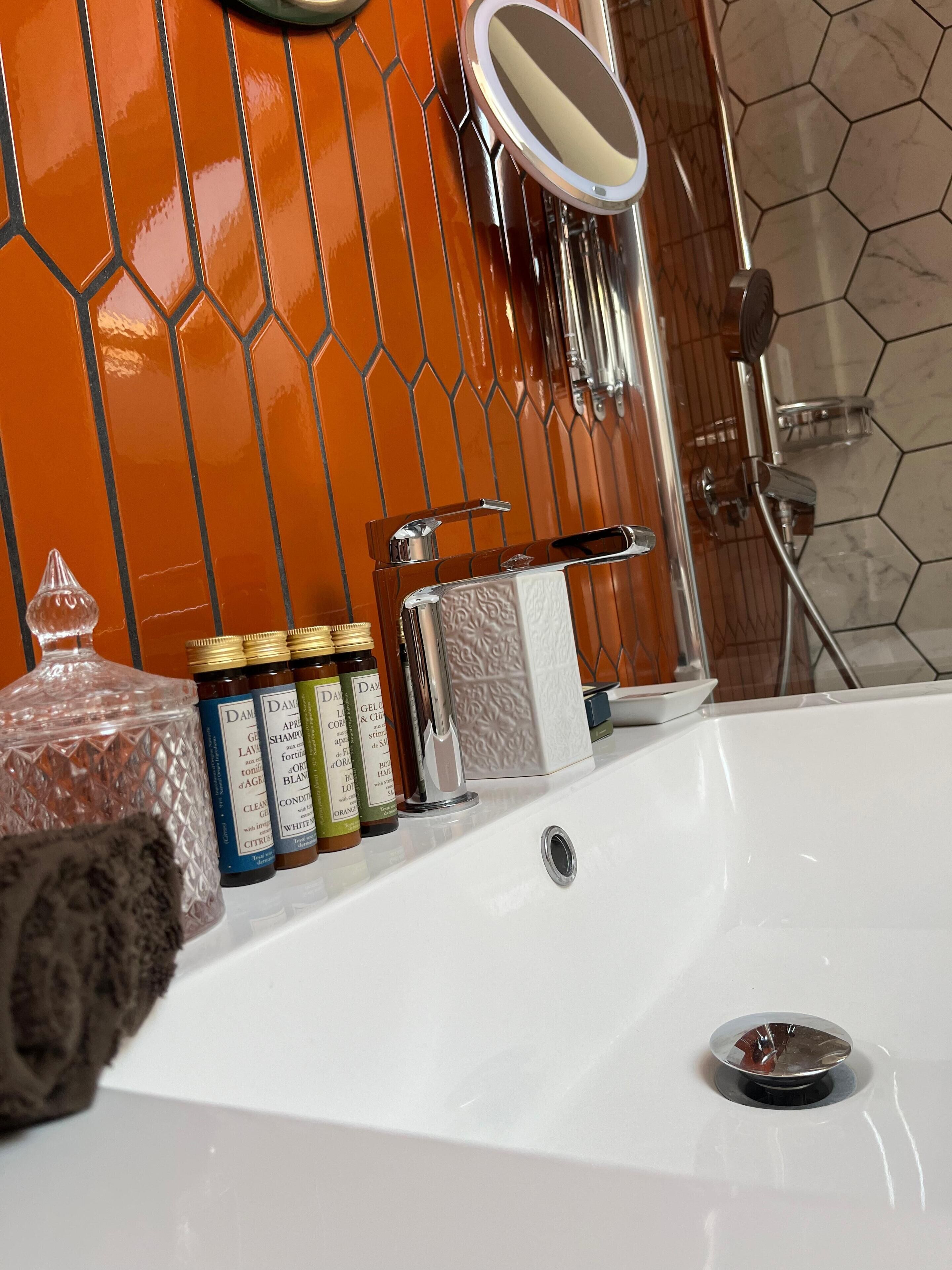 A bathroom with a white sink, a mirror, and a shower. There are bottles of shampoo and conditioner on the sink.